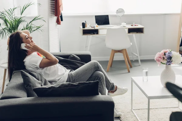 Vue latérale de la jeune femme transgenre souriante parlant par téléphone sur le canapé à la maison — Photo de stock