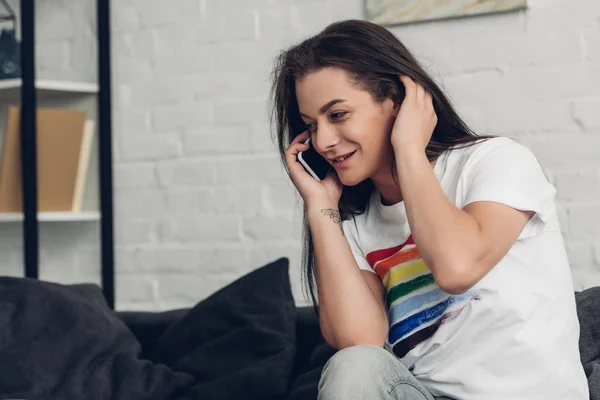 Улыбающаяся молодая трансгендерная женщина разговаривает по телефону на диване дома — стоковое фото