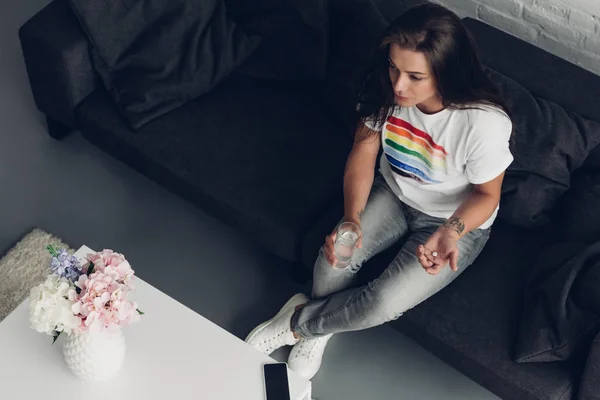 Vue grand angle de la jeune femme transgenre avec pilule et verre d'eau assis sur le canapé — Photo de stock