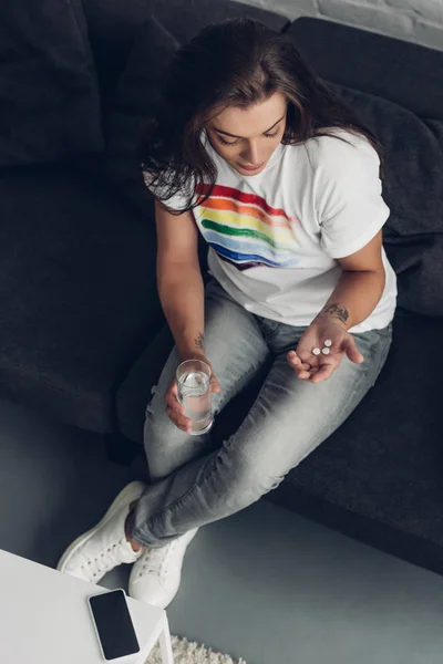 Vue grand angle de jeune femme transgenre déprimée avec des pilules et un verre d'eau assis sur le canapé — Photo de stock
