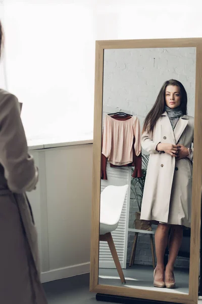 Young transgender woman in trench coat looking at mirror at home — Stock Photo