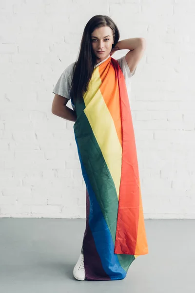 Atraente jovem transgênero mulher cobrindo com bandeira do orgulho e olhando para a câmera na frente da parede de tijolo branco — Fotografia de Stock