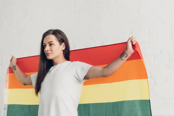 Mujer transgénero sosteniendo bandera de orgullo frente a la pared de ladrillo blanco - foto de stock