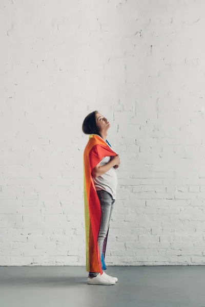 Young transgender woman covering shoulders with pride flag in front of white brick wall — Stock Photo