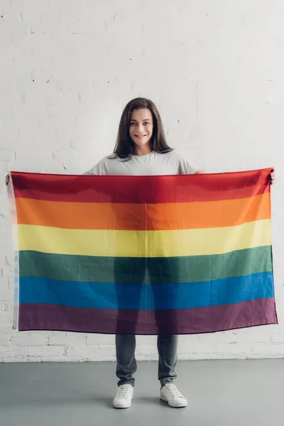 Mujer transgénero feliz sosteniendo la bandera de orgullo frente a la pared de ladrillo blanco - foto de stock