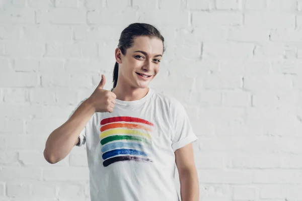Felice donna transgender in t-shirt bianca con bandiera orgoglio mostrando pollice davanti al muro di mattoni bianchi — Foto stock