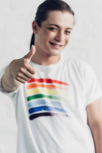 Молодая трансгендерная женщина в белой футболке с флагом гордости, показывающая большой палец перед стеной из белого кирпича — стоковое фото