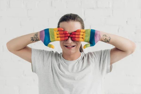 Mujer transexual sonriente cubriendo los ojos con las manos pintadas en colores de la bandera del orgullo delante de la pared de ladrillo blanco - foto de stock
