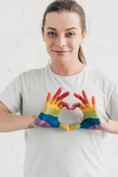 Sonriente joven transexual mujer haciendo signo de corazón con las manos en colores de la bandera de orgullo delante de la pared de ladrillo blanco - foto de stock