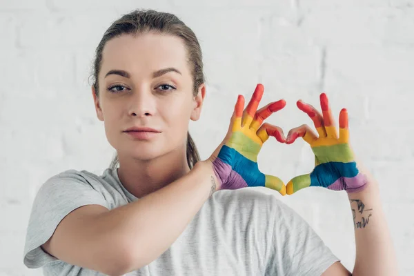 Giovane donna transgender che fa segno di cuore con le mani nei colori della bandiera orgoglio davanti al muro di mattoni bianchi — Foto stock