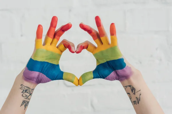Tiro recortado de las manos pintadas en colores de la bandera del orgullo que muestra el gesto del corazón delante de la pared de ladrillo blanco - foto de stock