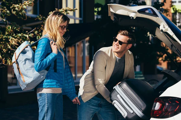 Homme mettre des bagages dans la voiture avec petite amie souriante à proximité — Photo de stock