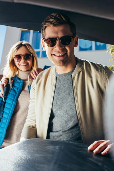Sonriente hombre poniendo equipaje en el coche mientras novia de pie detrás en la calle - foto de stock