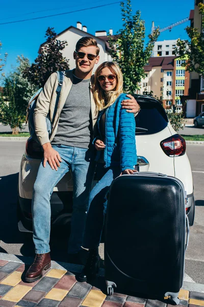 Пара улыбающихся туристов с багажом рядом с машиной на улице — стоковое фото
