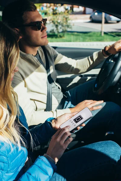 Mujer usando el teléfono inteligente con el logotipo uber en la pantalla mientras el marido conduce el coche - foto de stock