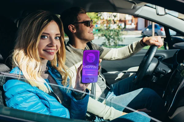 Улыбающаяся женщина, показывающая смартфон с надписью во время вождения автомобиля мужем — стоковое фото