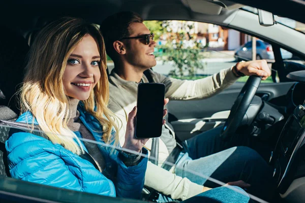 Улыбающаяся женщина показывает смартфон с пустым экраном во время вождения автомобиля бойфрендом — стоковое фото