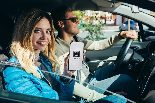 Улыбающаяся женщина показывает смартфон с логотипом Uber на экране во время вождения автомобиля мужем — стоковое фото