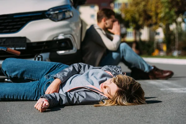 Selective focus of injured woman lying on road after car accident with shocked car driver behind — Stock Photo