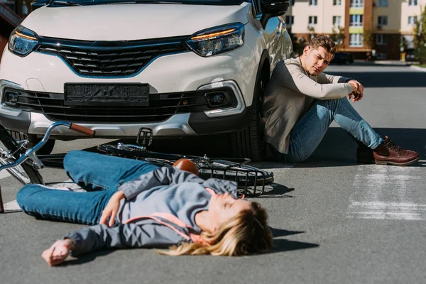 Selective focus of injured woman lying on road after car accident with car driver behind — Stock Photo