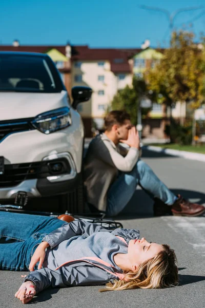 Selective focus of injured woman lying on road after car accident with shocked car driver behind — Stock Photo