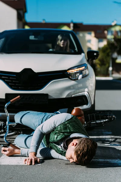 Woman sitting in car and looking at injured cyclist lying on road after traffic collision — Stock Photo