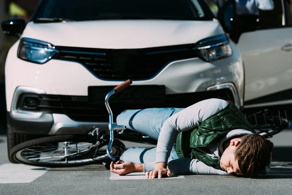 Injured young cyclist lying with bicycle on road after motor vehicle collision — Stock Photo