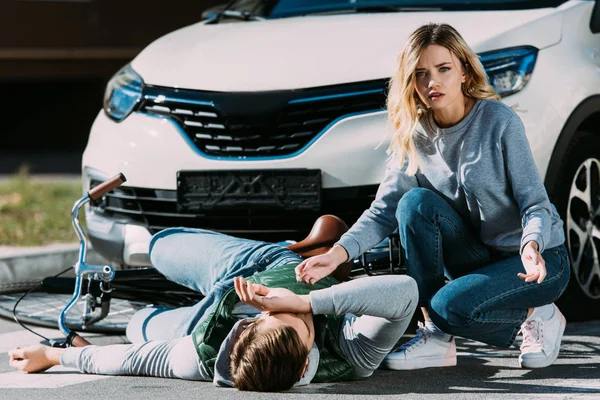 Shocked young woman looking at camera while injured cyclist lying with bicycle on road after traffic collision — Stock Photo