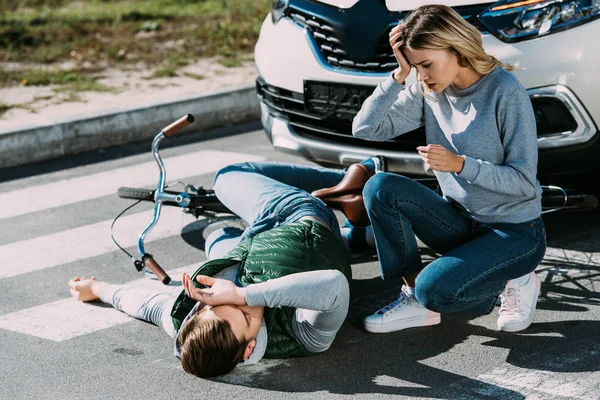 Shocked young woman looking at injured cyclist lying with bicycle at car accident — Stock Photo