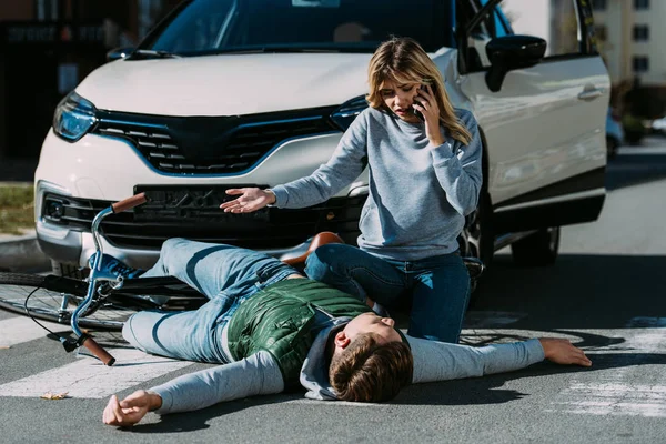 Mujer llamando de emergencia y mirando a ciclista lesionado acostado en la carretera después de la colisión de tráfico - foto de stock