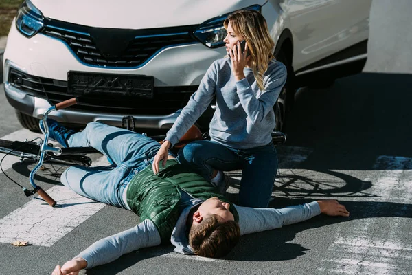 Vista de ángulo alto de la mujer llamando a la emergencia ciclista blanco lesionado acostado en la carretera después de la colisión de tráfico - foto de stock