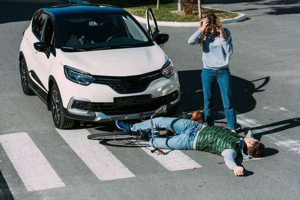 Vista de ángulo alto de la mujer de pie cerca de ciclista lesionado después de accidente de coche - foto de stock