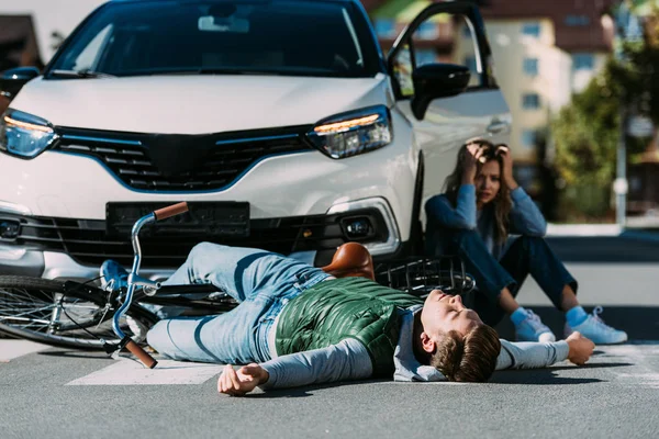 Ciclista lesionado acostado en la carretera y la mujer sentada cerca de coche después de la colisión de tráfico - foto de stock
