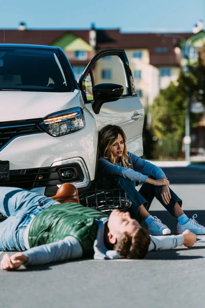 Jovem ciclista ferido deitado na estrada e mulher sentado perto do carro após colisão veículo a motor — Fotografia de Stock