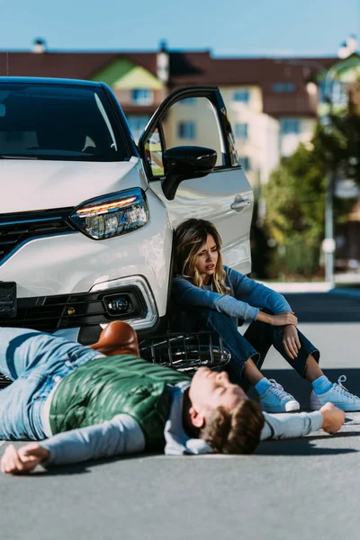 Injured young cyclist lying on road and woman sitting near car after motor vehicle collision — Stock Photo