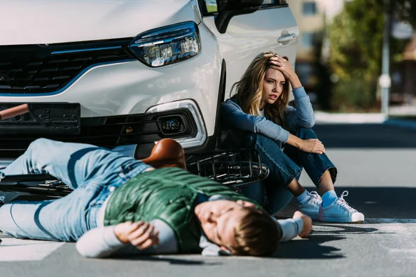 Mujer sentada cerca de coche mientras ciclista lesionado acostado en la carretera después de la colisión de tráfico - foto de stock