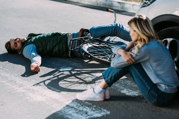 Conductor de coche sentado en la carretera y mirando ciclista lesionado después de accidente de coche - foto de stock