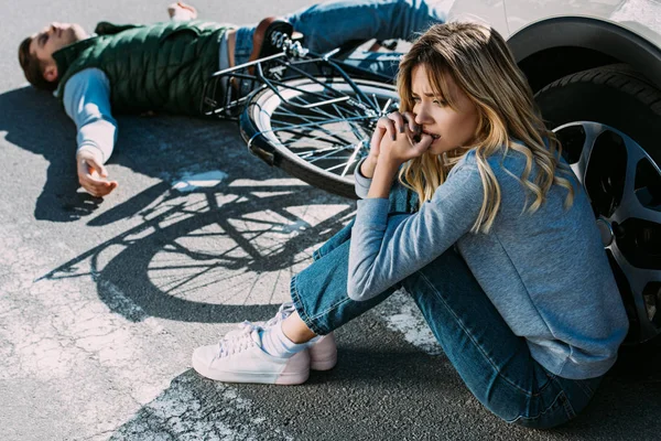 Asustada joven sentada cerca de coche después mientras ciclista acostado en la carretera después de la colisión de tráfico — Stock Photo