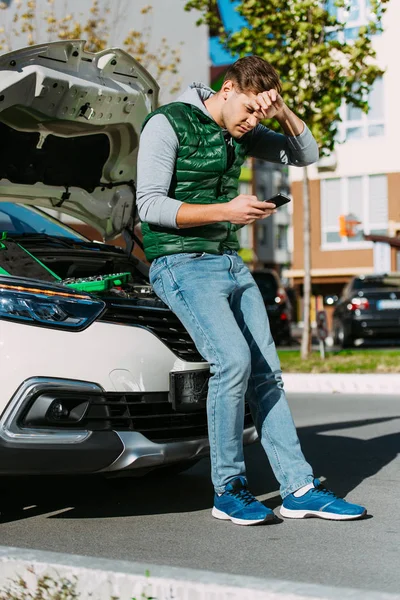 Verärgerter junger Mann sitzt auf kaputtem Auto und benutzt Smartphone — Stockfoto