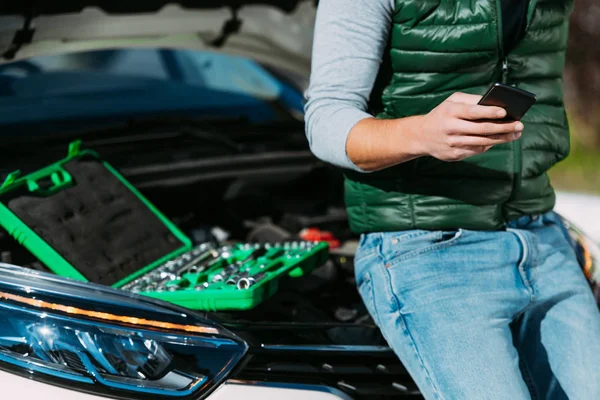 Cropped shot of man sitting on broken car and using smartphone — Stock Photo