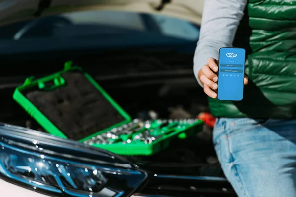 Cropped shot of man holding smartphone with skype app while sitting on broken car with toolbox — Stock Photo