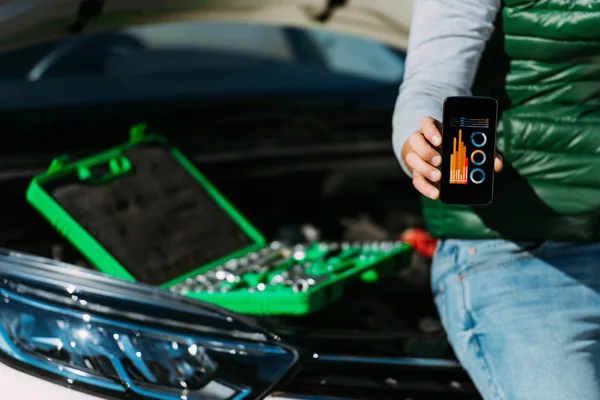 Ritagliato colpo di uomo in possesso di smartphone con grafici sullo schermo mentre seduto su un'auto rotta con cassetta degli attrezzi — Foto stock