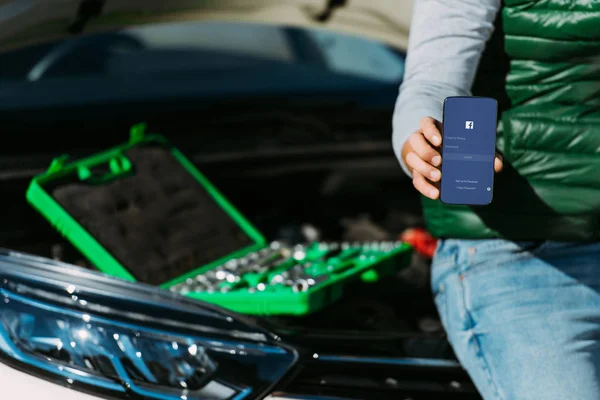 Schnappschuss von Mann, der Smartphone mit Facebook-App hält, während er mit Werkzeugkiste auf kaputtem Auto sitzt — Stockfoto