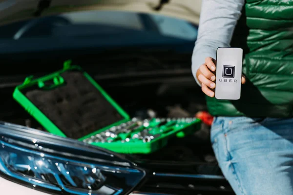 Tiro recortado de homem segurando smartphone com aplicativo uber enquanto sentado em carro quebrado com caixa de ferramentas — Fotografia de Stock