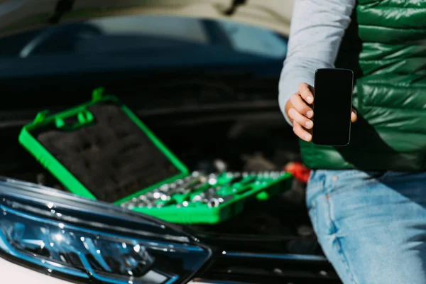 Cropped shot of man holding smartphone with blank screen while sitting on broken car with toolbox — Stock Photo