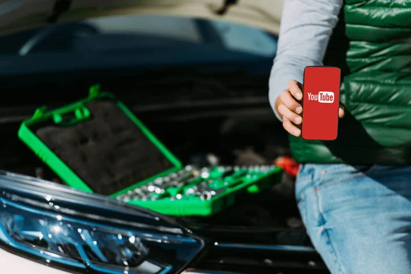 Plan recadré de l'homme tenant smartphone avec application youtube tout en étant assis sur la voiture cassée avec boîte à outils — Photo de stock