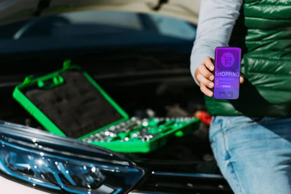 Schnappschuss von Mann, der Smartphone mit Shopping-App in der Hand hält, während er mit Werkzeugkiste auf kaputtem Auto sitzt — Stockfoto