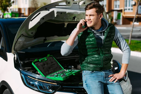 Улыбающийся молодой человек разговаривает по смартфону, сидя на сломанной машине — стоковое фото