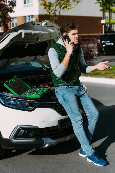 Junger Mann spricht mit Smartphone, während er auf kaputtem Auto sitzt — Stockfoto