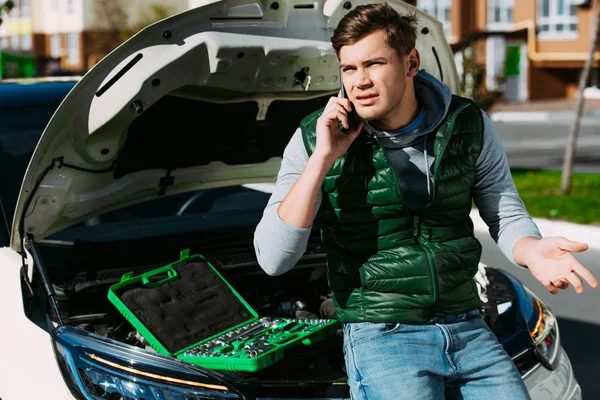 Emotionaler junger Mann spricht mit Smartphone, während er auf kaputtem Auto sitzt — Stockfoto